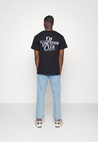 Czarna bawełniana koszulka z dużym białym napisem "ON VACATION CLUB" na plecach. W połączeniu z jasnoniebieskimi dżinsami oraz wielokolorowymi trampkami.