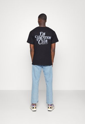 T-shirt en coton noir avec le texte blanc "ON VACATION CLUB" en grand au dos. Associé à un jean en denim bleu clair et des baskets multicolores.