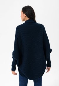 Tumedat sinine ülemõõduline kudumis-sviiter ribatud tekstuuriga ja kõrge kaelusega, pikad varrukad ja kumer hemline.