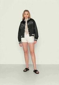 Jaqueta bomber preta com letras brancas, blusa em padrão zebra bege, calções brancos e sandálias pretas. Tecido suave, corte descontraído, design desportivo.