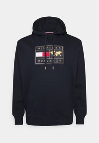 Mörkblå huvtröja med en frontgrafik i guld och flera färger, som visar en världskarta, logotekst och dragskoaccenter. Mjukt tyg, avslappnad passform.