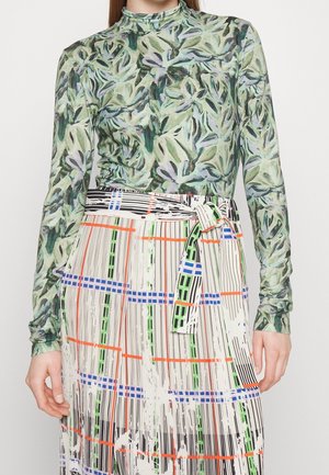 Femme portant un haut manches longues vert à motif feuilles et une jupe taille haute avec des motifs multicolores en grille et rayures.