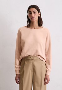 Lichtroze sweatshirt met lange mouwen en een ronde halslijn. Zachte textuur en een subtiele merklogo in het midden. Gecombineerd met beige broek.