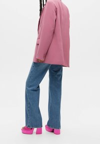 Blazer rose, jean évasé bleu et sandales à plateforme rose vif. Le blazer est doté de deux boutons, le jean présente une texture usée.