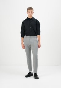 Jonge man die staat, gekleed in een zwart overhemd met knopen, grijze slim-fit broek, witte sokken en zwarte gepolijste kostuum schoenen op een effen witte achtergrond.