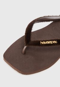 Rudos rudi sandalai su tekstūruota guma ir pūkuotu rudu dirželiu, ant kurio yra auksinis "havaianas" logotipas.