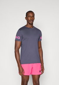 Tumši zils īsām piedurknēm T-krekls ar rozā zīmolu uz piedurknēm, kombinēts ar rozā šortiem ar melniem akcentiem. Vienkāršs, moderns dizains.