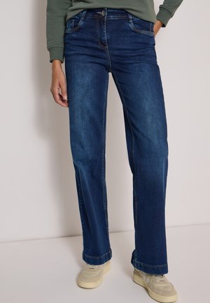 Personne portant un jean droit bleu foncé, des baskets beige clair et un haut vert à manches longues, debout devant un fond uni clair.
