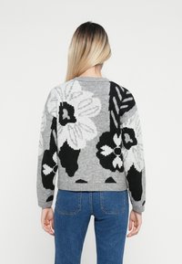 Maglione grigio lavorato a maglia con grandi motivi floreali in bianco e nero, scollatura rotonda e polsini e orlo a coste. Indossato con jeans blu.