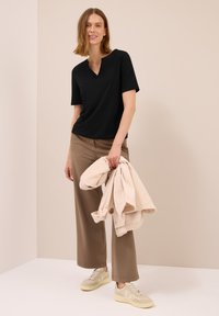 Frau steht an einer beigen Wand, trägt ein schwarzes kurzärmliges Oberteil, braune weit geschnittene Hose, hellbeige Sneakers und hält eine beige Jacke.