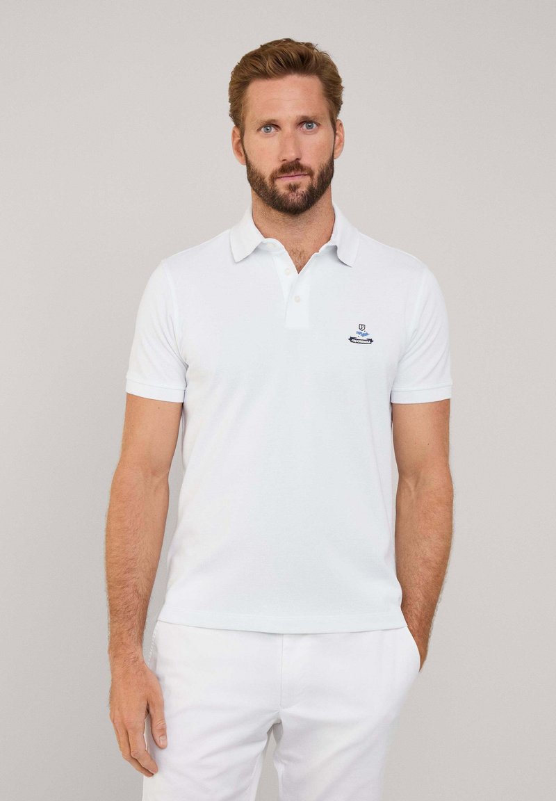 Camisa polo blanca con cuello y mangas cortas. Presenta un pequeño logo azul en el lado izquierdo del pecho. Combinada con pantalones blancos.