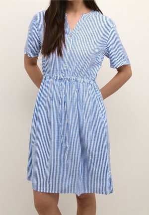 Kobieta ubrana w niebiesko-białą sukienkę w pionowe paski sięgającą kolan, z krótkimi rękawami, zapinaną na guziki z przodu, z wiązaniem w talii.