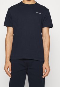 Mężczyzna w prostym, ciemnoniebieskim t-shircie marki Calvin Klein i pasujących niebieskich spodniach, stojący z rękami swobodnie opuszczonymi wzdłuż ciała.