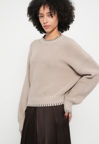 Bēša adīta džemperis ar apaļu kakla izgriezumu, ribojamām aprocēm un apakšmalu, ar smalkām tumšām detaļām apkakles malā. Brīvs piegriezums, teksturēta auduma.