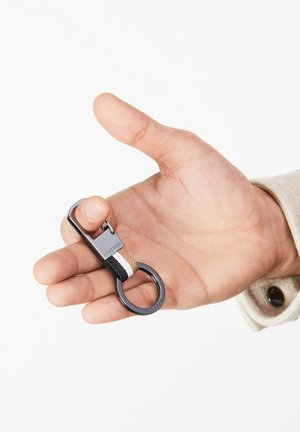 Keychain in hand, featuring metallic clip, black and beige woven strap with white accents, circular ring, sleek design, and branding.