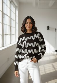 Kobieta o włosach do ramion, ubrana w czarny sweter z białymi falistymi paskami oraz białe spodnie, stojąca w oświetlonym słońcem pokoju.