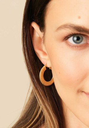 Gold textured hoop earring worn on a woman’s left ear with brown hair and light skin against a beige background.