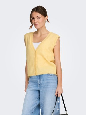 Žena v lehkém žlutém bezrukávovém cardiganu přes bílý top a světle modré džíny, držící černou kabelku na bílém pozadí.