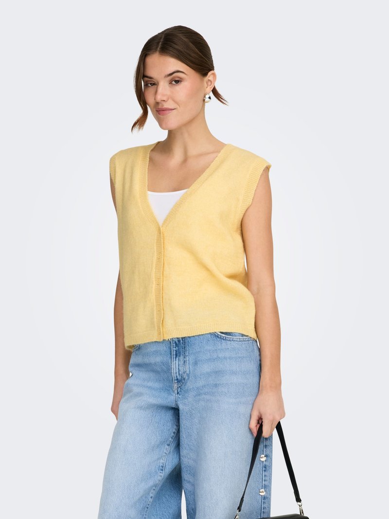 Ženska, ki nosi svetlo rumen brezrokavnik čez belo majico in svetlo modre kavbojke, drži črno torbico na belem ozadju.