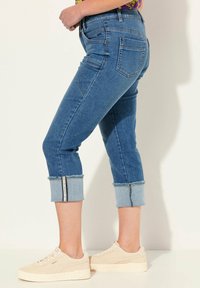 Modré džínové kalhoty s ohrnutými nohavicemi, které mají světle modrý lem. Perfektně padnou s pěti kapsami a výrazným švem. Spárované s béžovými teniskami.