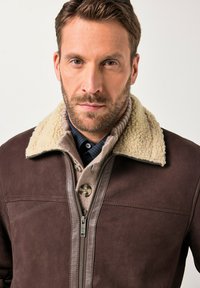 Hombre con cabello corto castaño y barba, vistiendo una chaqueta de ante marrón con cuello de borreguillo crema sobre un suéter abotonado y una camisa oscura.