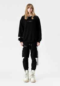 Sudadera negra de gran tamaño con una etiqueta frontal, combinada con pantalones jogger negros que presentan detalles desgastados y gráficos, completado con botas blancas.