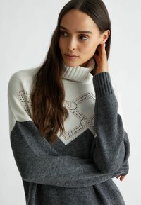 Maglione a collo alto grigio e bianco lavorato a maglia con design geometrico e decorazioni. Presenta polsini a coste e una vestibilità ampia. Texture morbida.