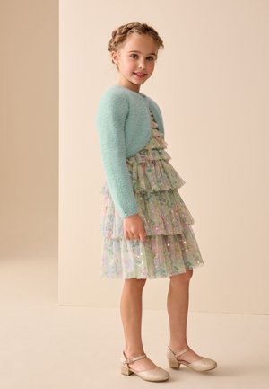 Joven niña sonriendo, lleva un vestido floral de capas con lentejuelas, un cárdigan azul claro y peludo, y zapatos Mary Jane dorados y brillantes.