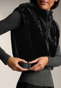 Vesta fără mâneci din blană artificială neagră, cu închidere prin fermoar, prezentând un top gri texturat cu mâneci lungi și o curea neagră cu prindere.