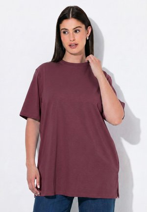 Frau mit langen dunklen Haaren, die ein übergroßes kurzärmeliges bordeauxrotes T-Shirt und blaue Jeans trägt, berührt den Ausschnitt und schaut zur Seite.