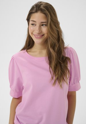 Jonge vrouw met lang golvend haar, draagt een lichtroze top met opgeblazen mouwen, lacht en kijkt opzij tegen een effen achtergrond.