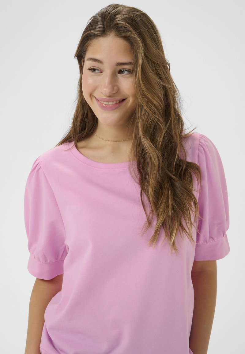 Młoda kobieta z długimi, falującymi włosami, ubrana w jasnoróżową bluzkę z bufkami, uśmiechająca się i patrząca w bok na jednolitym tle.