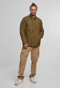 Pikowana oliwkowa kurtka z zatrzaskami i kieszenią na klatce piersiowej, zestawiona z jasnobrązowymi spodniami cargo i białymi trampkami.