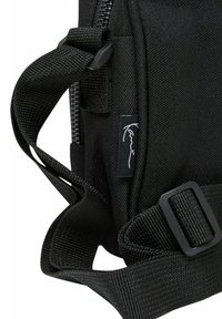 Sort stoftaske med struktureret materiale, justerbar strap og en logo-tag, med en lynlås synlig øverst.