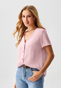 Lichtroze blouse met korte mouwen en een gestructureerde afwerking, met knopen aan de voorkant en een gegolfde halslijn, gecombineerd met een blauwe spijkerbroek en een bruine riem.