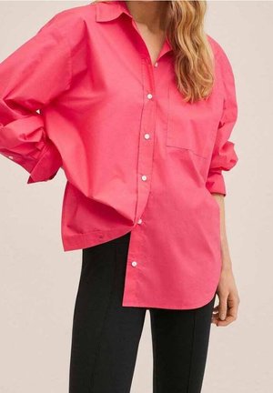 Kobieta ubrana w oversize'ową, jasnoróżową koszulę na guziki z kieszenią z przodu oraz czarne dopasowane spodnie na neutralnym tle.