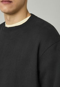 Sweatshirt noir surdimensionné avec un col rond côtelé et une texture douce. Superposé à un t-shirt de couleur claire. Design minimaliste, sans motifs.