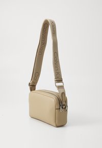 Beige skulderveske med en glatt tekstur, rektangulær form, to glidelåser, og et vevd reim med "Calvin Klein" merke.
