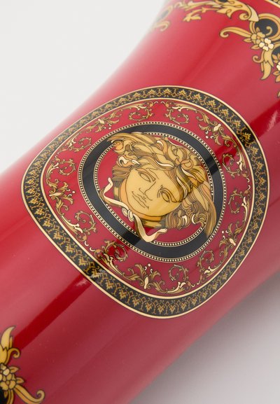 Czerwony wazon z misternym złoto-czarnym ornamentalnym wzorem, z centralnym medalionem przedstawiającym twarz, o gładkim, błyszczącym wykończeniu.