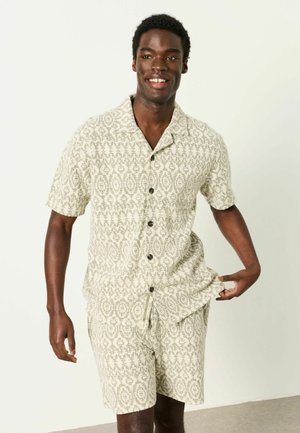 Lächelnder Mann, der ein beige gemustertes Kurzarm-Hemden mit Knöpfen und passende Shorts trägt, vor einem einfarbigen hellen Hintergrund stehend.