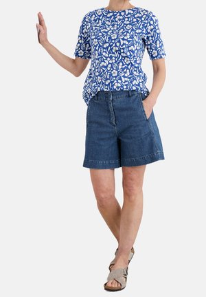Osoba ubrana w niebieską bluzkę z krótkim rękawem ozdobioną kwiatowym wzorem, ciemne dżinsowe szorty i beżowe sandały odkrywające palce, stojąca z uniesioną jedną dłonią.