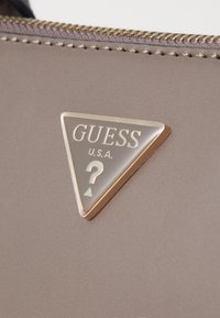 Logótipo triangular com "GUESS U.S.A." em dourado sobre um material de cor taupe, apresentando um ponto de interrogação prateado e um detalhe de costura distinto.
