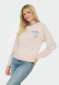 Sweatshirt rose clair avec col rond, coupe décontractée et manches longues. Présente un texte imprimé en bleu et un logo sur le devant.