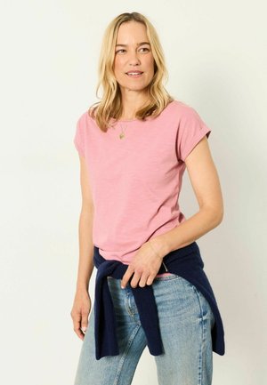Woman with blonde hair wearing a pink short-sleeve shirt, blue jeans, and a navy sweater tied around her waist, looking to the side.