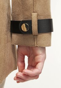 Jasnobrązowy zamszowy rękaw kurtki z czarnym skórzanym paskiem i złotym guzikiem, podkreślający teksturę i kontrast kolorów. Ręka spoczywa w pobliżu.