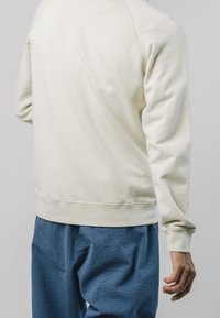 Persoon in een crème kleurige sweatshirt met lange mouwen en een textuurbroek in het blauw, staande tegen een effen lichte achtergrond.