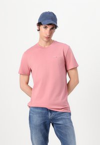 Camiseta de algodón rosa con mangas cortas y escote redondo, que presenta un pequeño logotipo. Llevada con jeans de mezclilla azul y una gorra de béisbol azul.