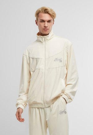 Krämfärgad zip-jacka i mjukt material med ribbade ärmslut och broderad logotyp på bröstet; matchad med byxor med elastiskt midjeband.