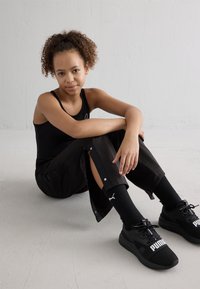 Camisola atlética preta e calças largas pretas com botões de lado, combinadas com ténis pretos da Puma e meias às riscas. O cabelo encaracolado está estilizado de forma natural.