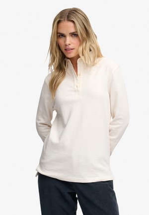 Kvinde med langt blondt hår, iført en cremefarvet langærmet poloshirt og mørke bukser, står med hænderne bag ryggen.